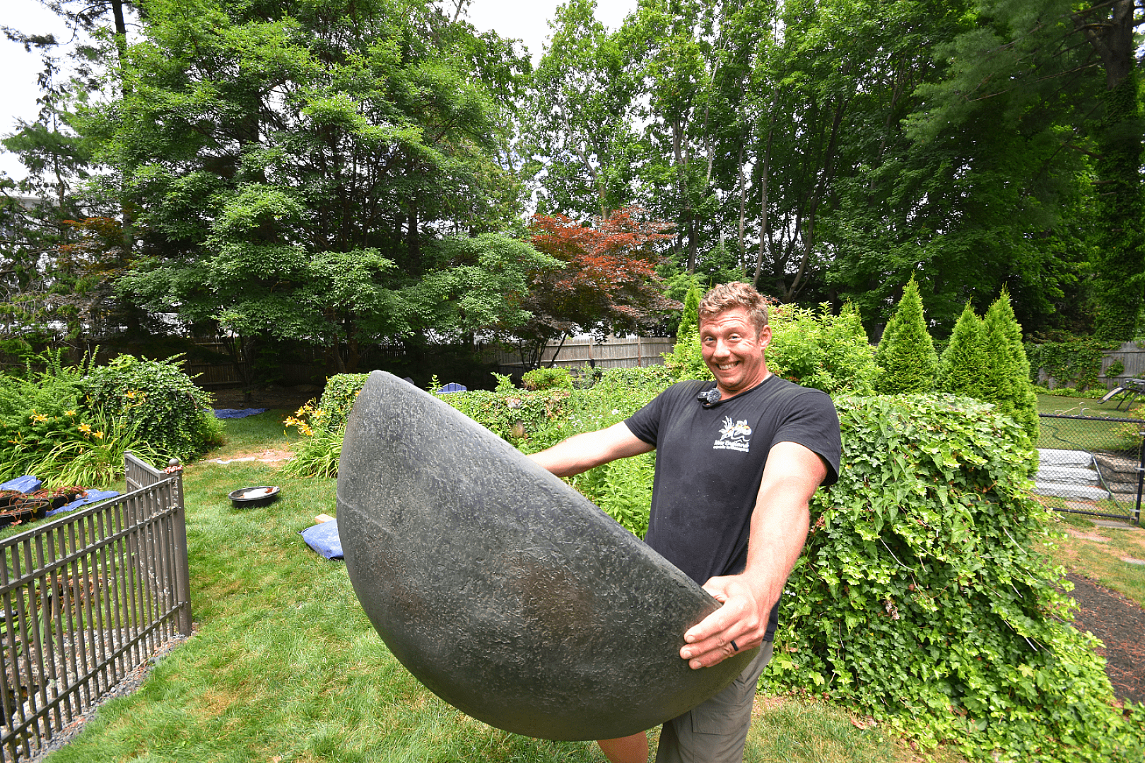 Smiling man holding large concrete half-sphere in garden.